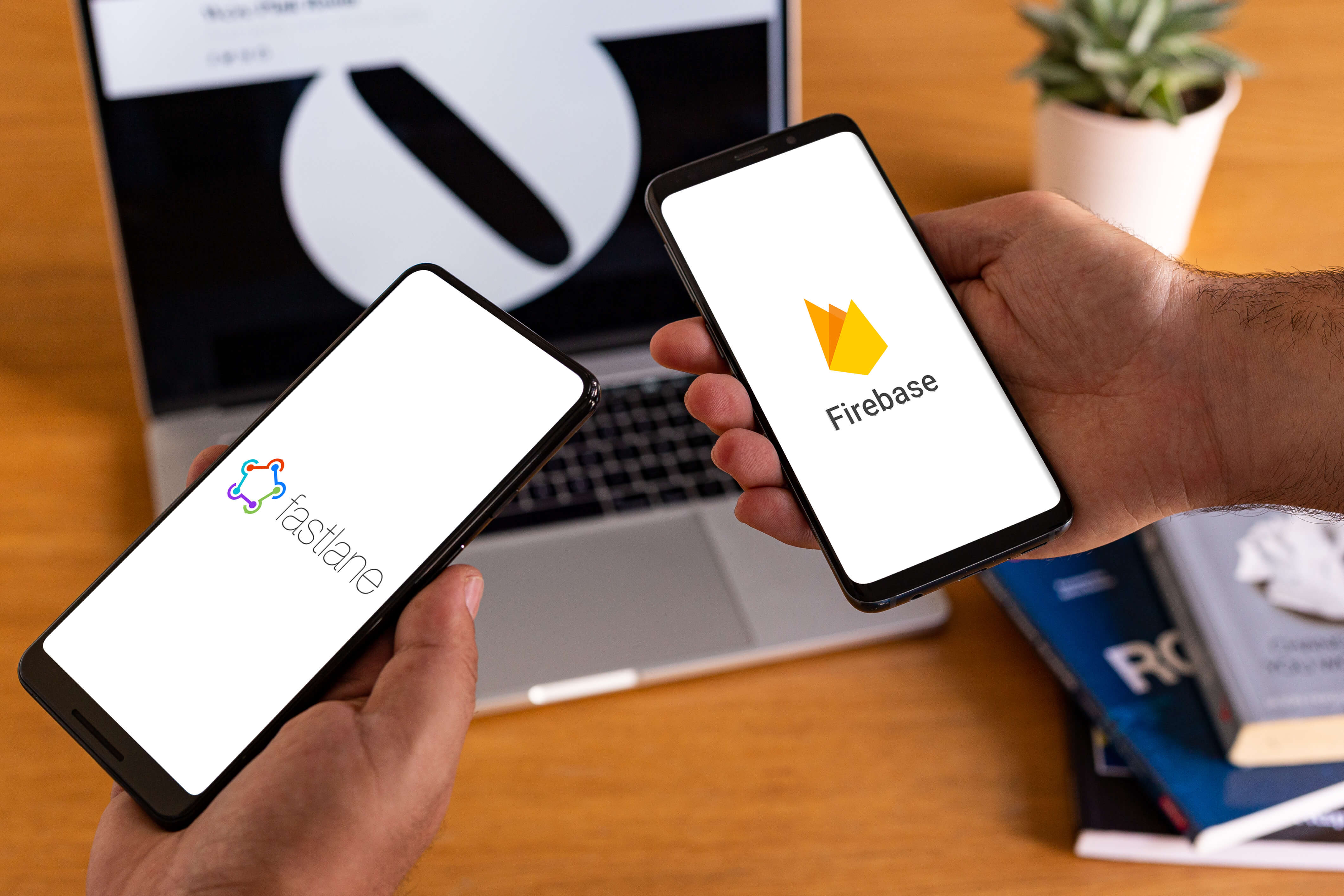 Two smartphones with the fastlane and firebase logos, with a mac laptop, books and a plant in the background